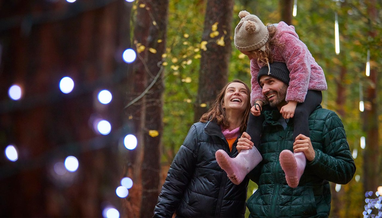 A family of 3 smile as they walk through twinkling lights in the forest. The little girl sits on her dad's shoulders.