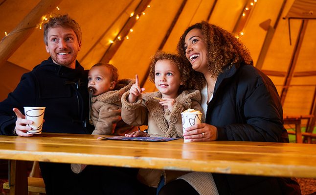young family sat in twinkling Tipi with hot drinks 