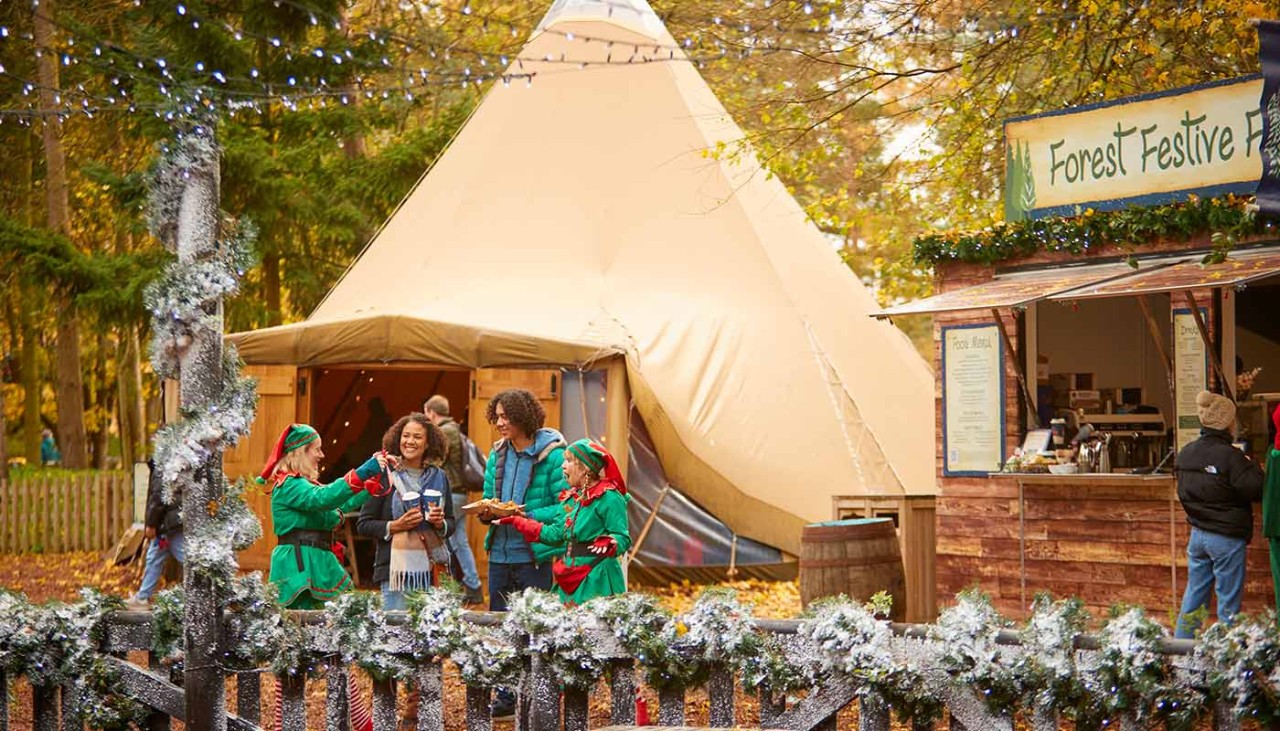 A couple standing with elves and eating food outside the Forest Festive Fayre Tipi.