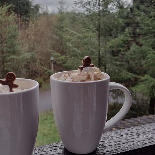Two mugs filled with cream and a gingerbread man poking out the top with forest views behind
