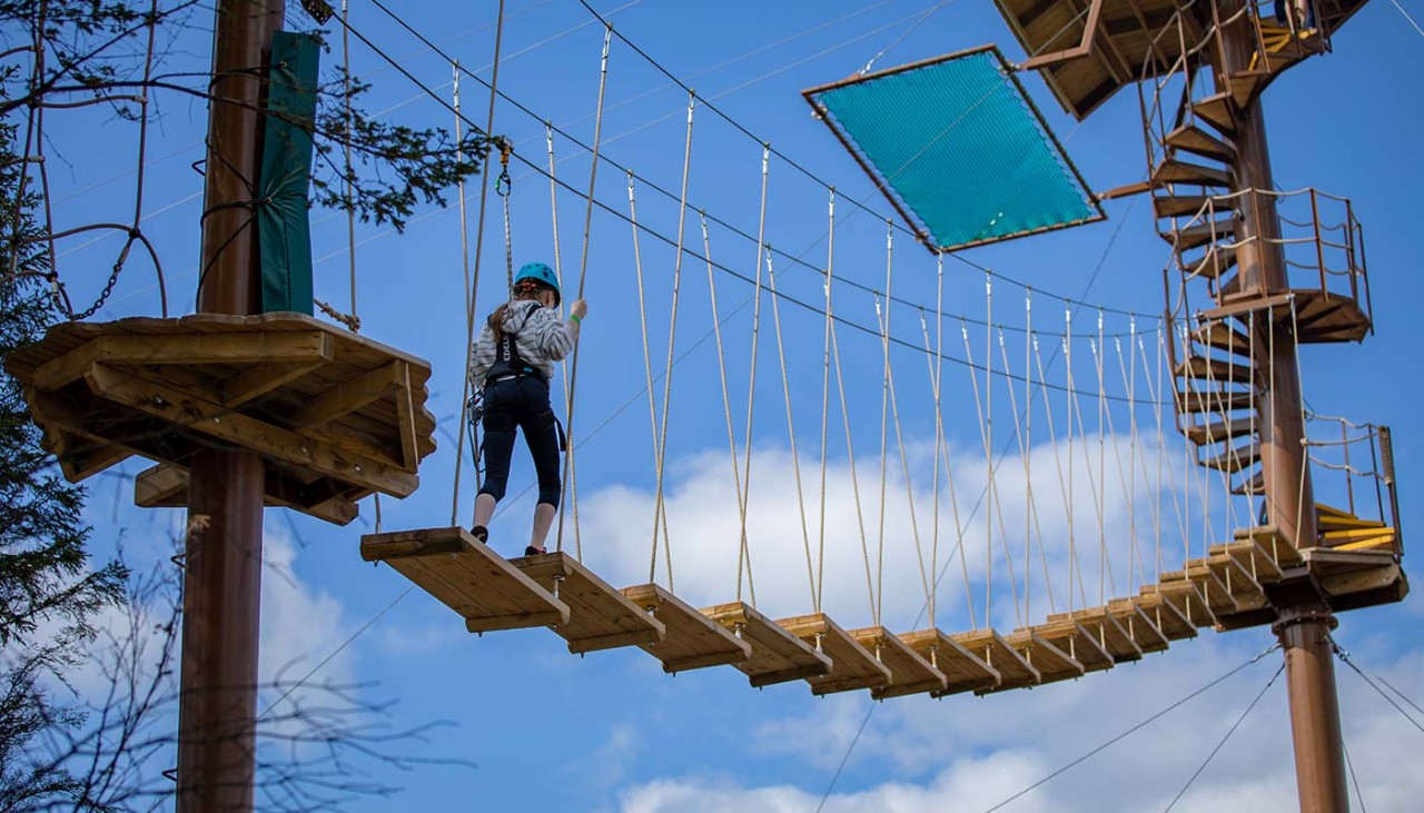 A girl making her way up to the top of the aerial adventure. A spiral staircase and zipline await her in the distance.