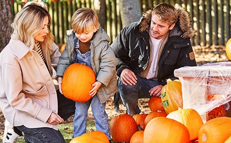 Two children hug a blue-and-white inflatable beach ball, smiling and splashing while wading chest-deep in a sunlit outdoor swimming pool, with blurred greenery in the background.