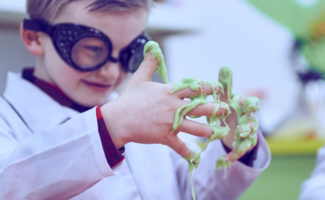 Child in oversized goggles manipulates gooey green slime, stretching it between sticky fingers, while wearing a white lab coat in a bright classroom or play-lab setting.