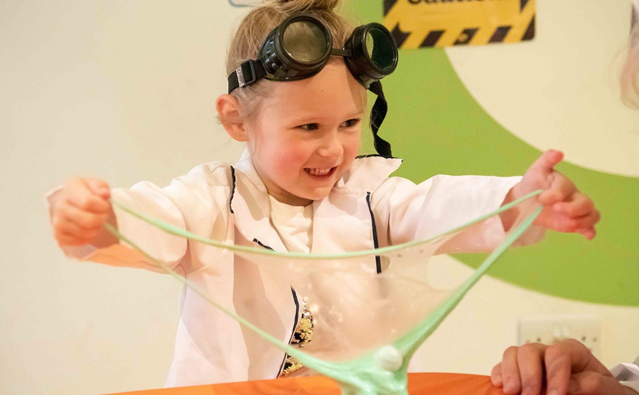 little girl smiling as she pulls green slime dressed in goggles and scientist jacket