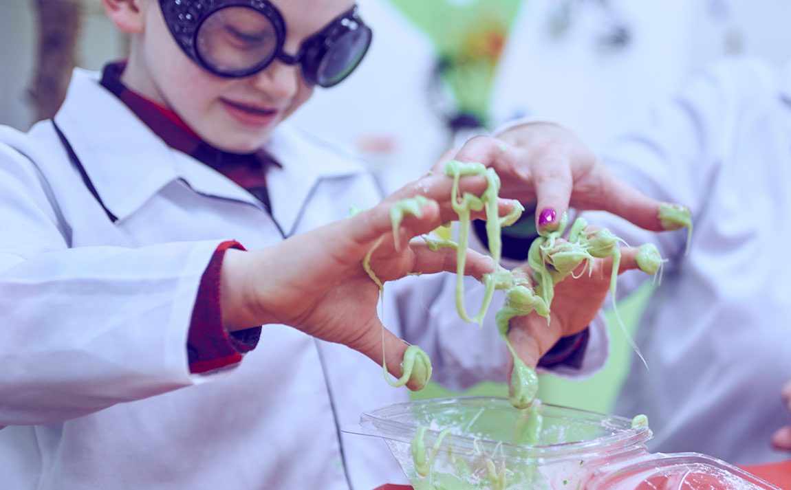young boy in scientist jacket and goggles sticking fingers into green slime