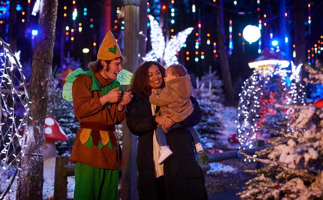 Mum holding baby in snowy and light-filled village with pixie entertaining the baby