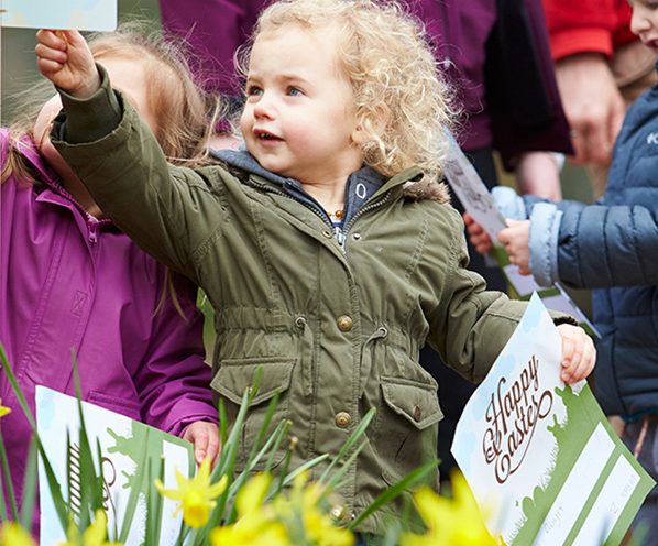A little girl holding an Easter leaflet standing near daffodils.