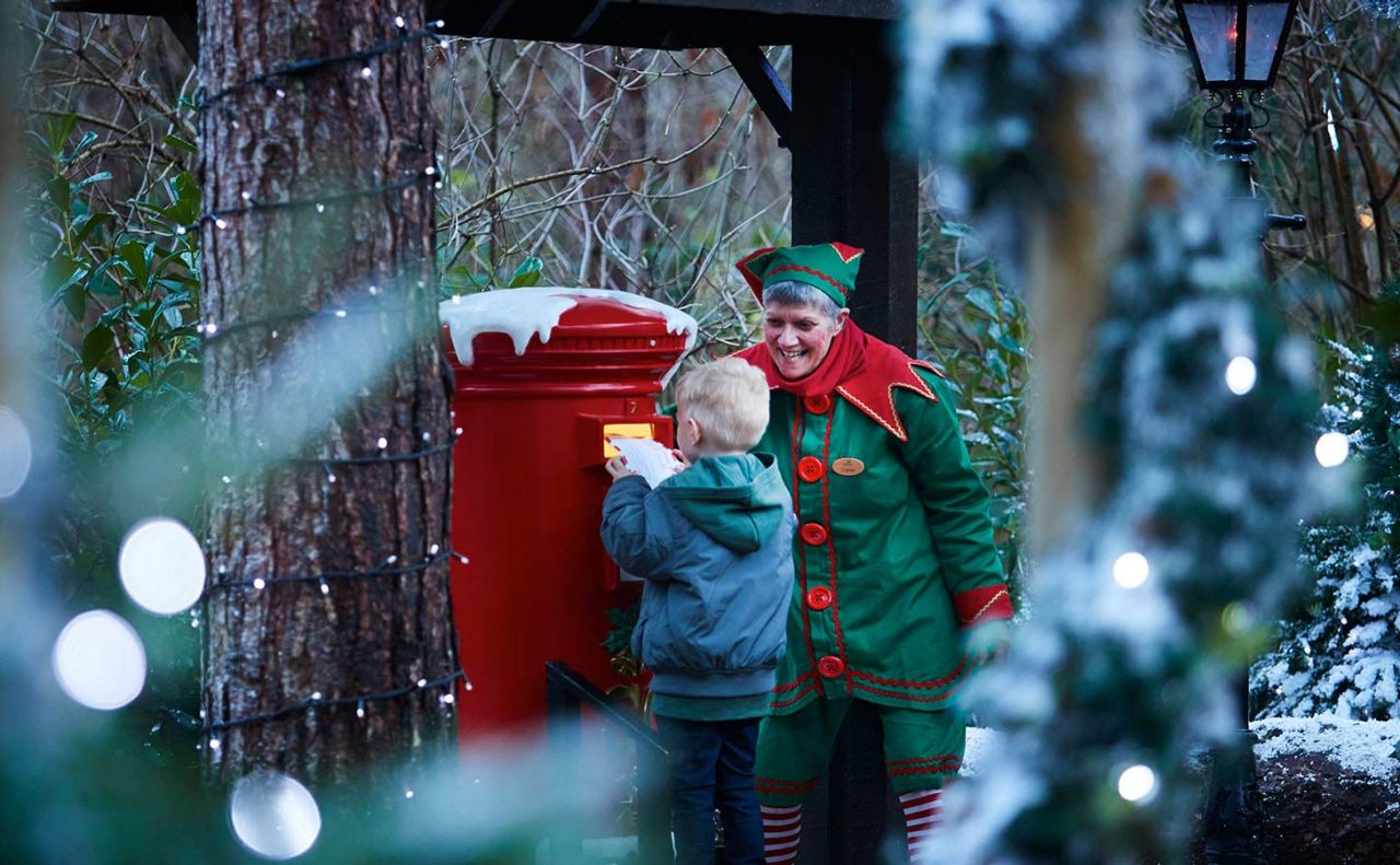 A child being encouraged by an elf posting a letter into a snow covered postbox 