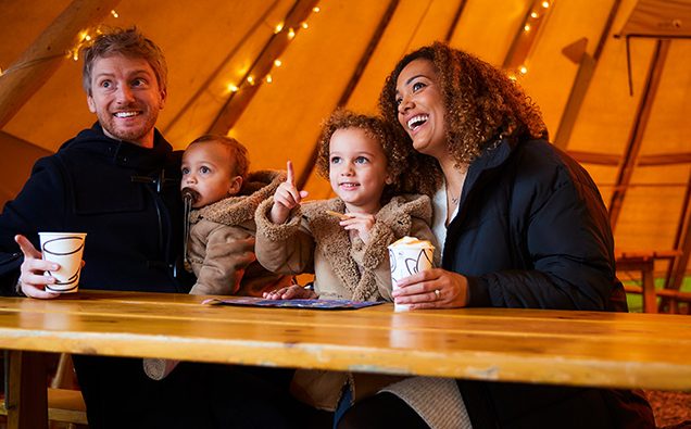 young family sat in twinkling Tipi with hot drinks 