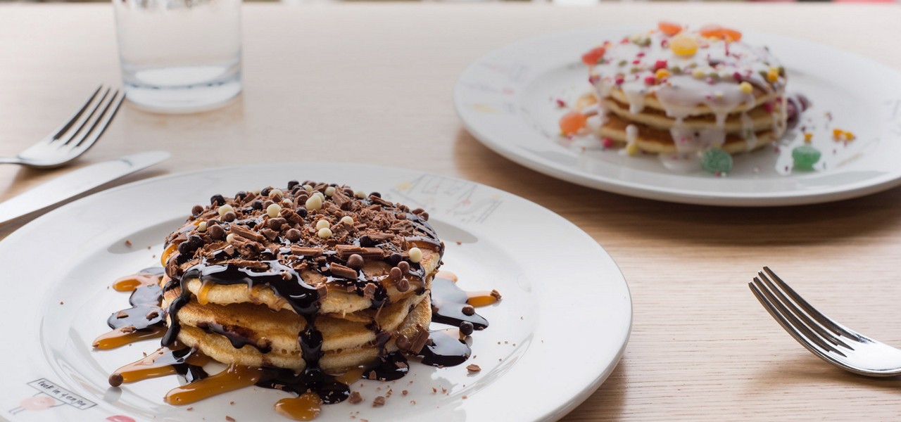 Stacked pancakes drizzled with chocolate and caramel sit on a plate, topped with chocolate shavings; another iced, candy‑topped stack sits behind. Forks and water glass rest on a wooden table.