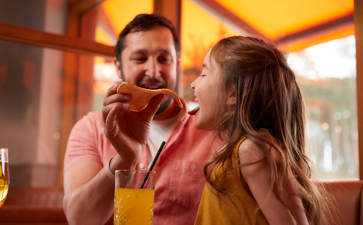 Dad feeding daughter a slice of pizza with her mouth open