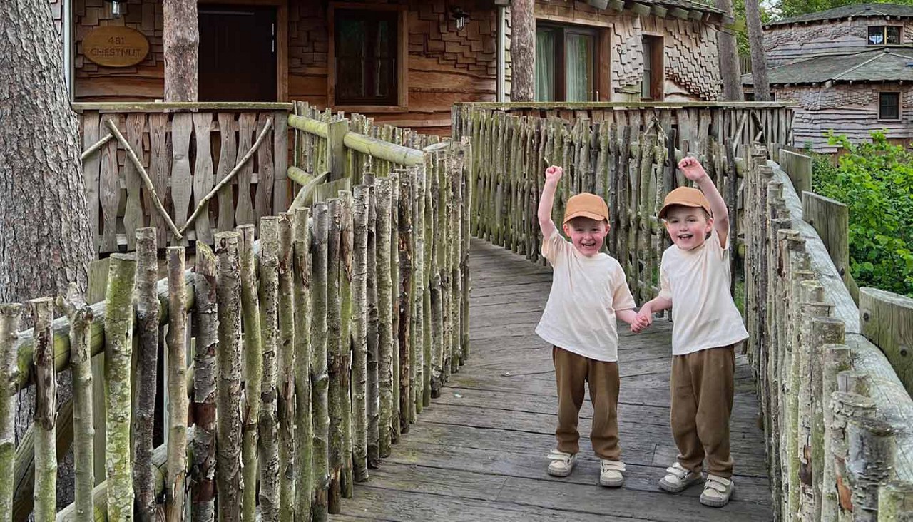 Two children hold hands and cheer, arms raised, while standing on a rustic wooden footbridge leading to treehouse cabins in a forest. Text: “CHESTNUT” and an unreadable number.