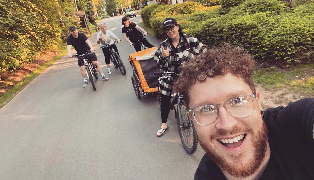 Cyclists ride together; front rider takes a smiling selfie while others follow, one towing an orange child trailer; scene unfolds on a quiet, tree-lined residential street in warm daylight.